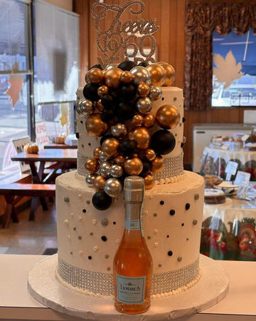 Decorative cake with gold, black, and silver balloons and a bottle of champagne on a table.