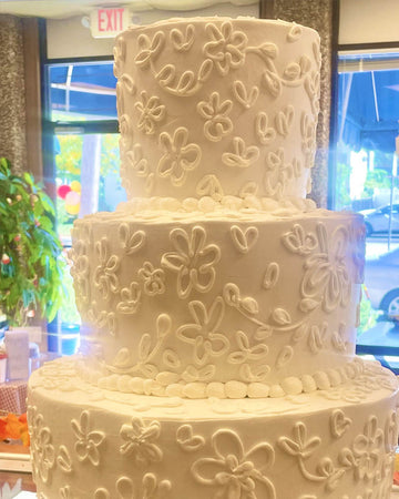 Three-tiered white wedding cake with floral patterns in a room with large windows.